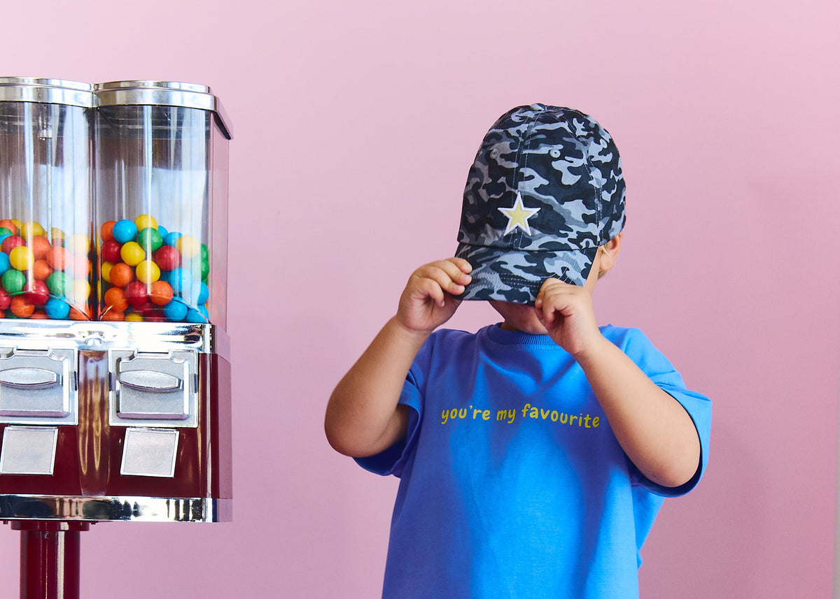 A toddler stands against a pink studio background wearing a blue t-shirt with yellow text, matching blue and black camouflage pants with yellow star accents, and a camouflage cap. A vintage-style ctwister machine with colorful gummy candies is visible on the left.