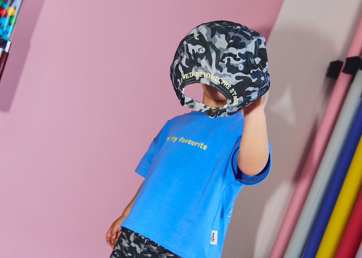 A child modeling a bright blue t-shirt, black and gray camouflage pants, and a matching camouflage cap, posed in a pink studio setting.