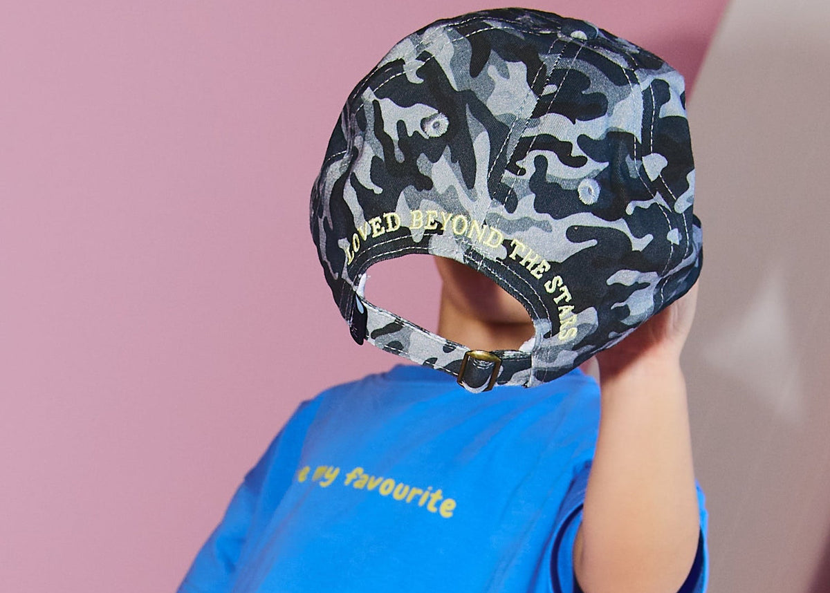 A child modeling a bright blue t-shirt, black and gray camouflage pants, and a matching camouflage cap, posed in a pink studio setting.